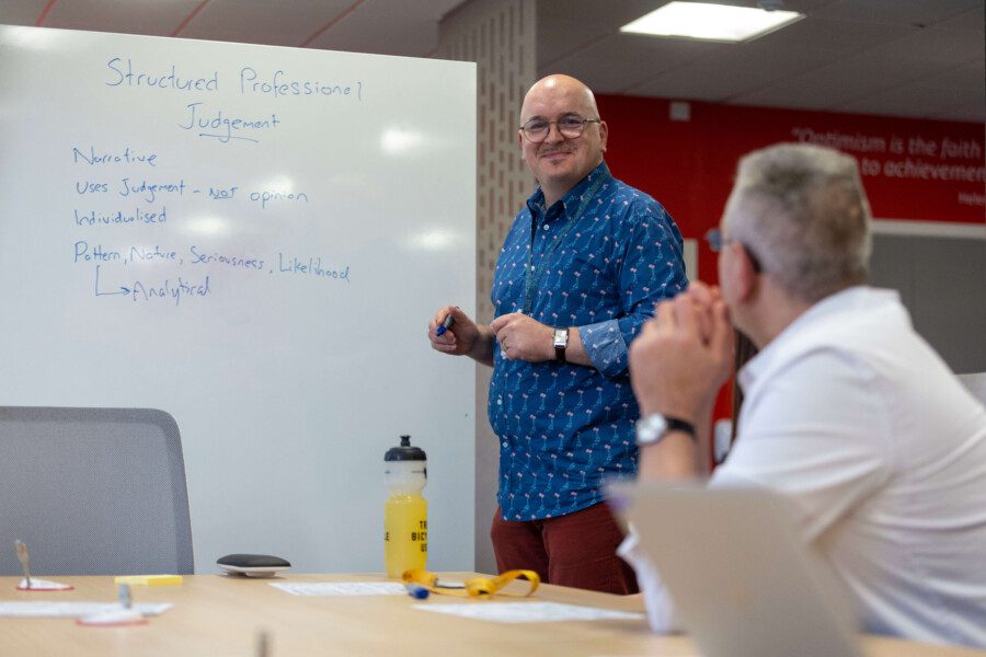 A classroom setting. One man is writing on a white board while another is watching.