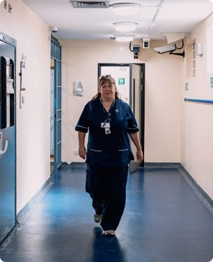 A nurse wearing scrubs walking down a corridor, looking into the camera