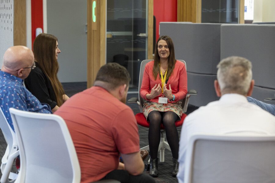 An office setting. Five people sitting on desk chairs are in a circle layout and in discussion