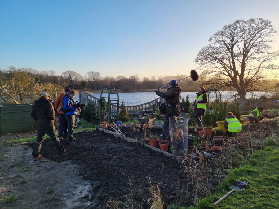 A behind-the-scenes photo of filming in a garden as the sun sets. There is a camera crew filming three people in high-vis vests.